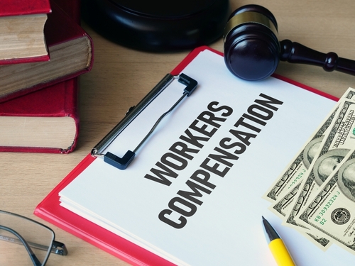 Clipboard labeled "Workers Compensation" with cash, a gavel, and law books on a desk.
